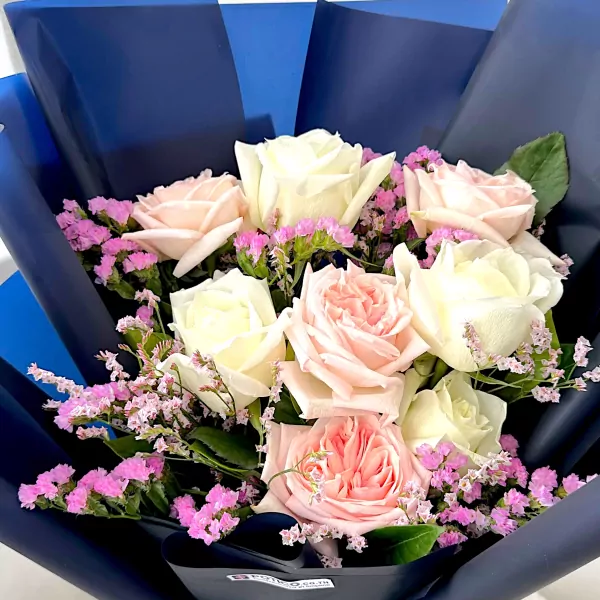 Bouquet of white and pink roses with small pink dried flowers, wrapped in dark blue paper.