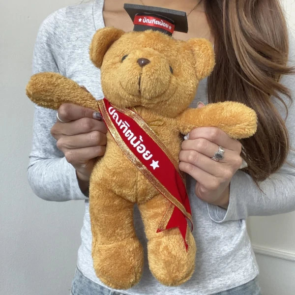 A teddy bear wears a graduation cap and a red sash with Thai writing.