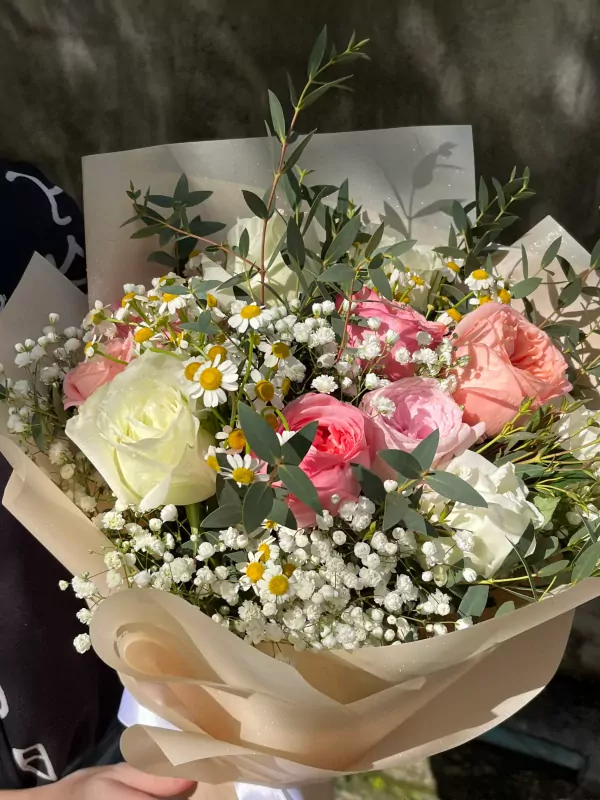A bouquet of pink and white roses with baby's breath, daisies, and eucalyptus in light brown paper.