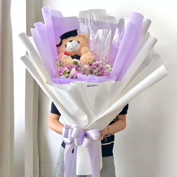 Graduation bouquet with teddy bear wearing a cap and dried flowers, wrapped in white and purple paper.