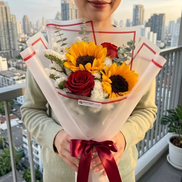 Person holding a bouquet of sunflowers, red roses, and white filler flowers, tied with a burgundy ribbon.