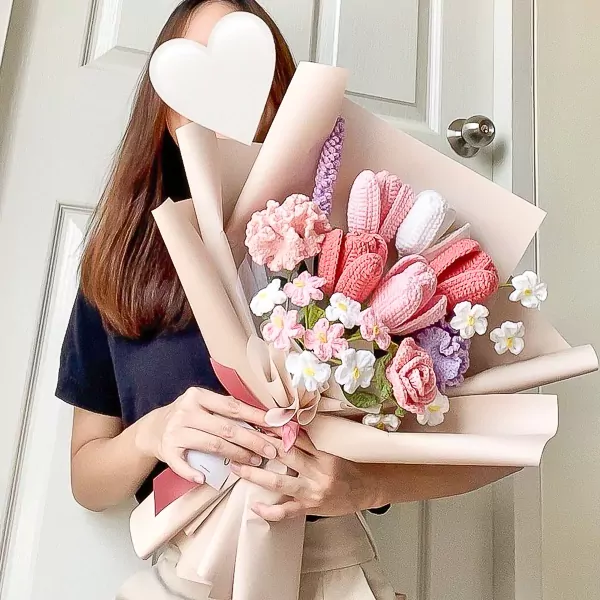 Crocheted pink tulips and carnations in a bouquet with green leaves, held by a person.