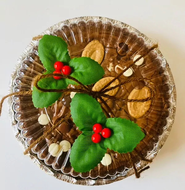 Decadent 6-inch Valrhona chocolate brownie topped with chocolate chips, white chocolate chunks, almonds, and festive holly sprigs in a foil pan.
