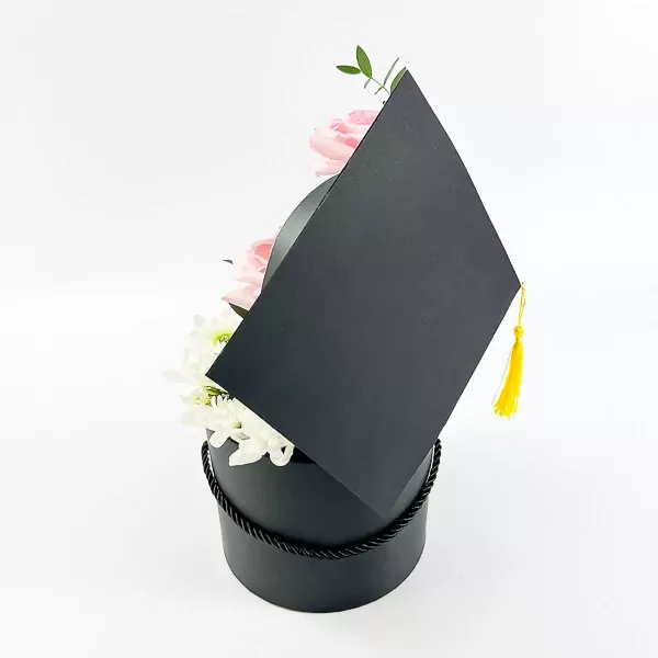 Black graduation hat box with pink roses and white mums, topped with a yellow tassel.