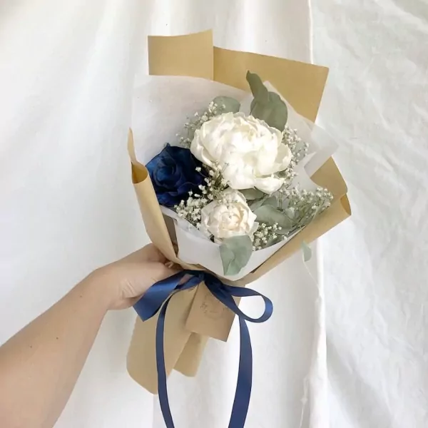 A bouquet of white and blue roses, peonies, and baby's breath, wrapped in brown paper and tied with a blue ribbon.
