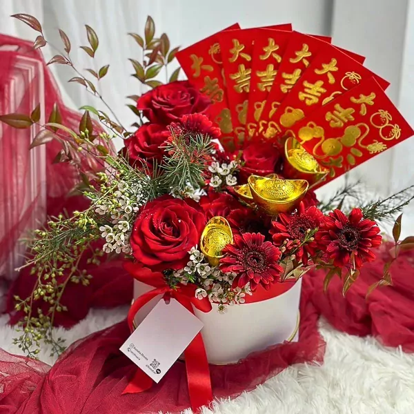 Red roses and gerberas in a white box adorned with red envelopes and gold ingots.