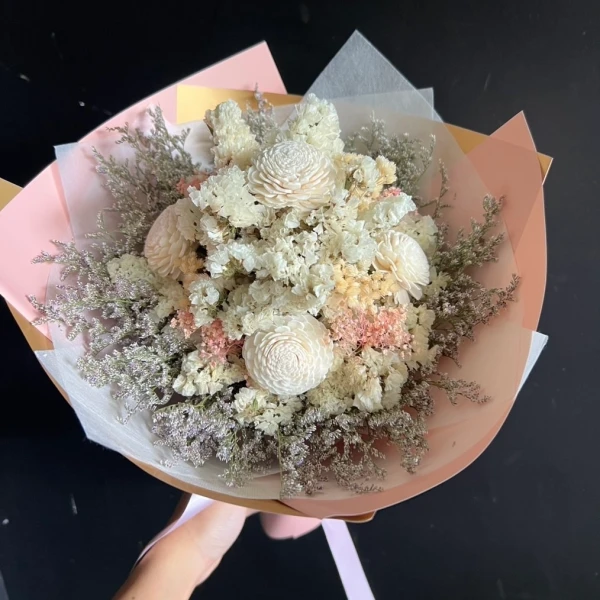 A dried floral bouquet with ivory blossoms and rustic foliage, wrapped in pink and gold paper.