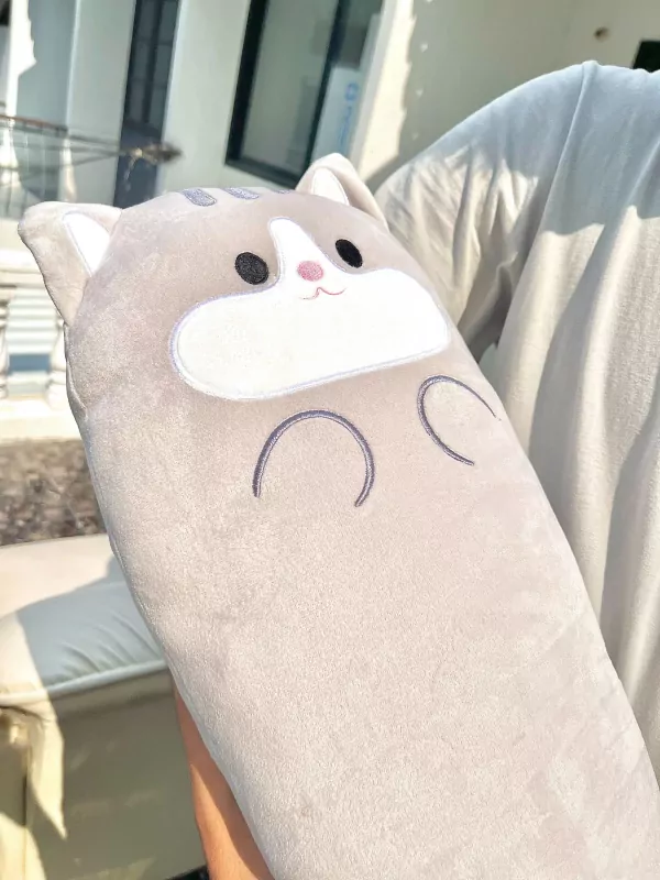 Plush grey cat pillow with embroidered face and paws, held by a person in a white t-shirt.