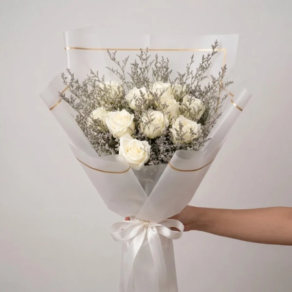 A hand holds a white roses bouquet wrapped in white paper with gold trim and a white ribbon.