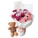 A teddy bear and a bouquet of pink and purple flowers in white wrapping, tied with a pink ribbon.