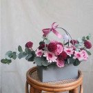Pink and purple floral arrangement in a grey box with a pink ribbon on a wicker stool.