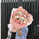 A bouquet of pink roses, carnations, blue star flowers, and white daisy-like flowers in pink paper.