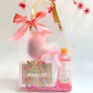A clear gift box with pink ribbon, a floral card, a pink pouch, and a cherry blossom body wash.
