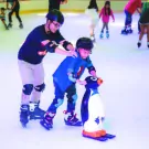 Man in helmet helps child in helmet on ice skates with penguin.