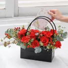 A hand places a floral card into a black box filled with red roses, hypericum berries, and eucalyptus.