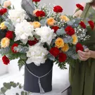 A luxurious bouquet of white peonies, red carnations, and orange roses in a dark grey box.