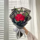 Red roses in black paper wrapping, tied with a white ribbon, held by a person's hand.