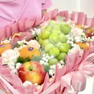 Fruit basket with green grapes, oranges, red apples, pink roses, and baby's breath.