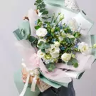 A bouquet of white lisianthus, hydrangea, and blue fillers with eucalyptus leaves, wrapped in light green and pink paper.