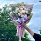 A bouquet of pale pink and white roses with eucalyptus, wrapped in purple and brown paper with a pink ribbon.