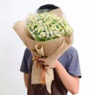 A person holds a bouquet of white daisies wrapped in burlap with a brown ribbon.