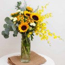 A tall glass vase filled with sunflowers, yellow orchids, and greenery.