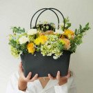 A black flower basket filled with yellow, white, and peach flowers, held by a person.