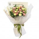 Bouquet of pink cappuccino roses and yellow carnations wrapped in white paper with a green ribbon.