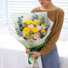 Woman in brown sweater holding a bouquet of yellow and pink roses with eucalyptus leaves.