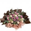 A bouquet of pink and white roses with purple accents, elegantly wrapped in brown and pink paper.