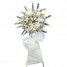 White floral wreath with orchids, roses, and greenery on a stand with a white bow.