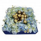 A blue floral arrangement with Ferrero Rocher chocolates and daisy accents in a dark blue box.