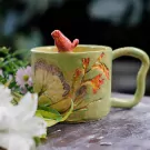 Green ceramic mug with sculpted butterfly and flowers, and a small red bird on the rim.