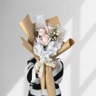 A bouquet of three light pink roses and baby's breath, wrapped in brown and white paper with a beige ribbon.