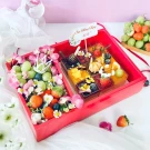 A red gift box filled with small cakes, fresh strawberries, blueberries, and grapes, decorated with flowers.