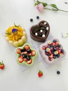 Four decorative fruit puddings, including mango, berry, and chocolate flavors, arranged on a white surface with scattered berries and a pink rose.
