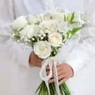 White wedding bouquet with roses, daffodils, and baby's breath, held by a person in a white shirt.