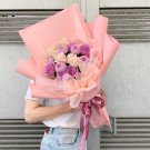 A bouquet of pink and white roses and purple chrysanthemums wrapped in pink paper, held by a person in a white t-shirt and jeans.