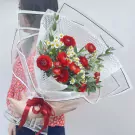 A woman holds a bouquet of red ranunculus and white tana flowers wrapped in clear and white paper, tied with a red ribbon.