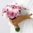 A bridal bouquet of purple and cream roses with small pink flowers, held by hands in a white dress.