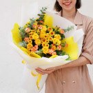 A woman holds a large bouquet of yellow roses, carnations, and daisies with yellow and white wrapping.