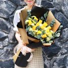 Woman holding a bouquet of yellow roses and baby's breath, wrapped in brown and black paper with a pink bow.