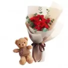 A teddy bear sits next to a bouquet of red roses and eucalyptus.