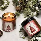 Two amber glass candles with wooden wicks sit in snow surrounded by pine branches, one lit.