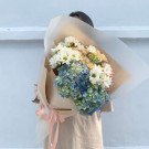 Bouquet of blue hydrangeas and white daisies wrapped in paper.