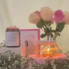 A jar of pink wax melts, a vase of pink and cream roses, and a lit oil diffuser on a pink display.