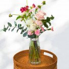 A bouquet of pink and white roses, alstroemeria, carnations, and eucalyptus in a clear glass vase on a woven tray.