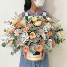 A person holds a round hat box filled with peach, pink, and cream roses, succulents, and eucalyptus.