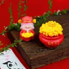 Golden Fortune Wax Set with a god of wealth figurine and a pot of gold, on a wooden stand with green sprigs.
