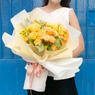 A woman in a white top holds a yellow and orange flower bouquet wrapped in pale yellow and white paper, tied with a pink ribbon.